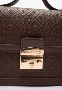 Sac à main marron texturé avec un motif en diamant et des accents floraux en relief. Présente un fermoir en ton doré marqué "ANNA FIELD". Matériel en cuir lisse.