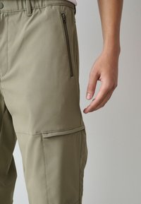 Kaki kravas bikses ar gludu tekstūru, ar sānu kabatu ar rāvējslēdzēju un ērtu piegriezumu. Roka sniedzas pret audumu.