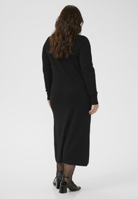 Robe noire à manches longues en matière douce, avec une coupe droite et une longueur mi-mollet. Portée avec des chaussures à talons à lanières noires.