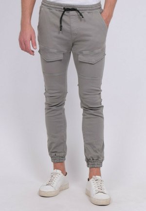 Homme portant un pantalon de jogging gris clair avec des poches cargo et un cordon de serrage noir, assorti à des baskets blanches sur un fond clair.