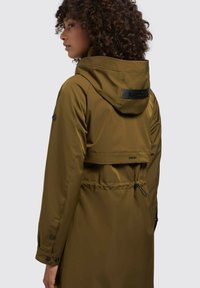 Giacca impermeabile verde oliva con vita regolabile tramite coulisse, cappuccio e dettagli con logo. Presenta polsini con bottone a pressione e un design elegante.