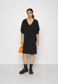 Robe noire en tricot avec un décolleté en V profond et des manches surdimensionnées. Assortie à des bottes noires à semelles épaises et à un petit sac orange.