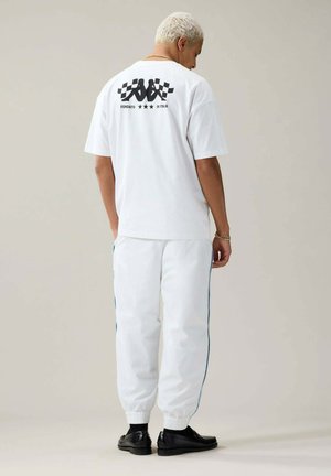 Persona con cabello corto y rubio que lleva pantalones blancos y una camiseta blanca con un logo negro y texto en la espalda, de pie sobre un fondo liso.