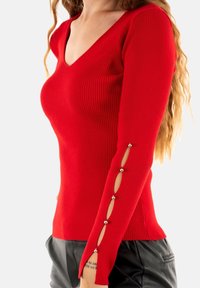 Vrouw in een aangesloten rode geribbelde trui met zilveren knopen en ovale uitsparingen aan de mouwen, gecombineerd met zwarte leren broek.