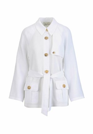 Femme portant une veste blanche boutonnée avec des poches et un pantalon à jambes larges, tenant une pochette tissée beige, accessoirisée de lunettes de soleil noires et de bracelets blancs.