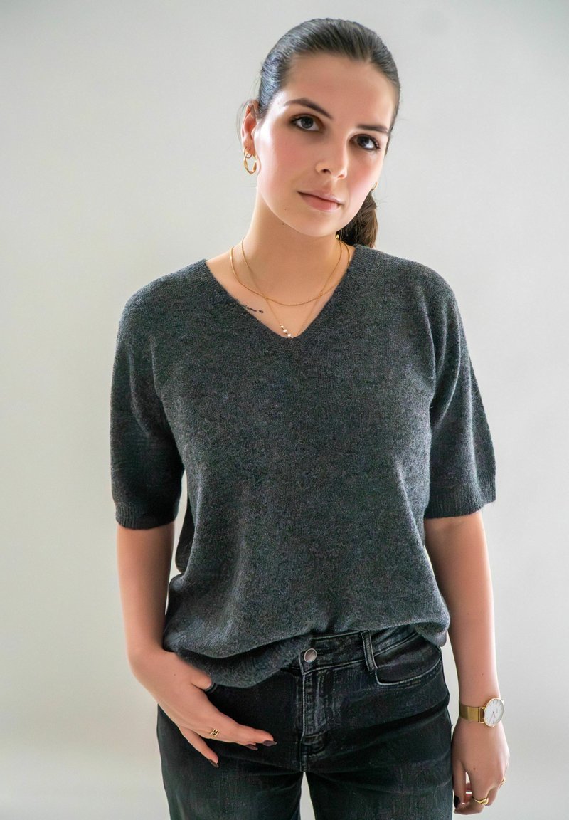 Grijze, kortemouwen v-hals trui van zacht materiaal; gecombineerd met donkere jeans. Geaccessoireerd met gouden sieraden en een horloge.