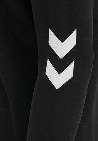 Svart långärmad skjorta med en mjuk yta och vit chevron-design på vänster ärm, som visar en minimalistisk estetik.