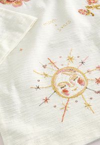 Tela bordada en blanco con un diseño de sol que presenta acentos en rosa y amarillo. Incluye elementos florales y el texto "luna y estrellas".