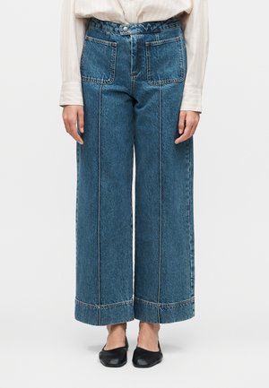 Kobieta w niebieskich jeansach z wysokim stanem i szerokimi nogawkami z przednimi naszywanymi kieszeniami oraz czarnych płaskich butach, stojąca na jednolitym tle.