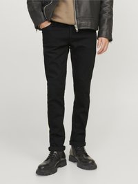Homme portant un jean noir retroussé, des bottes en cuir noires, une chemise marron et une veste en cuir noir avec une main dans la poche.