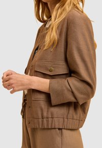 Femme portant une veste marron avec des poches à rabat à l'avant et des manches retroussées sur un pantalon assorti, devant un fond uni.