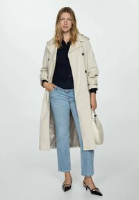 Beige trenchcoat met dubbelzijdige knopen, gecombineerd met een zwarte top en lichtblauwe jeans. Zwarte puntige schoenen en een witte handtas.