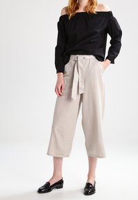 Blusa nera off-the-shoulder con maniche lunghe, abbinata a pantaloni a palazzo beige chiaro con vita legata. Scarpe nere lucide.