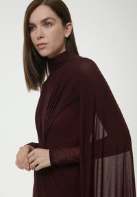Burgunderfarbene Robe mit hohem Kragen, plissiertem Oberteil und durchsichtigen Akzenten. Lange Ärmel und fließendes Design schaffen eine elegante Silhouette.