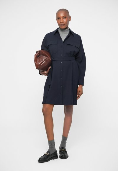 Μπλε πουκαμίσα φόρεμα με δύο μπροστινές τσέπες, κλείσιμο με κουμπιά, και ρυθμιζόμενη ζώνη. Συνδυασμένο με γκρι κάλτσες και μαύρες λοφάκρους, κρατώντας μια καφέ τσάντα.