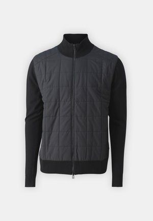 Melns zip-up vest ar quiltēta priekšpusi un adītas piedurknes. Iezīmē ribotas aproces un augstu apkakli, izgatavots galvenokārt no poliestera un vilnas.