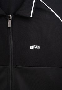 Svart zip-up jacka med en texturerad yta, med vita accentrandar och "UNFAIR" broderad i vitt på bröstet.