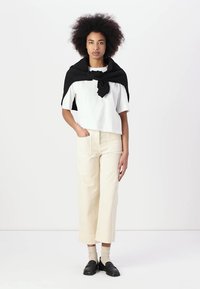 T-shirt bianco corto, cardigan nero appoggiato sulle spalle, pantaloni larghi color crema, calzini beige, mocassini neri, con capelli ricci e texturizzati.