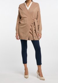 Cardigan avvolgente beige con maniche lunghe, dettaglio con cintura in vita e orlo a costine, abbinato a pantaloni fitted blu scuro e tacchi beige.