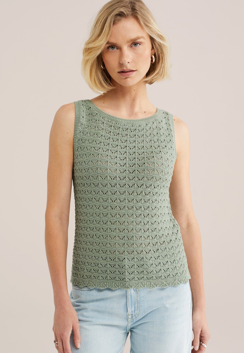 Femme aux cheveux blonds courts portant des boucles d'oreilles créoles dorées, un haut sans manches en tricot vert clair et un jean bleu clair sur un fond neutre.