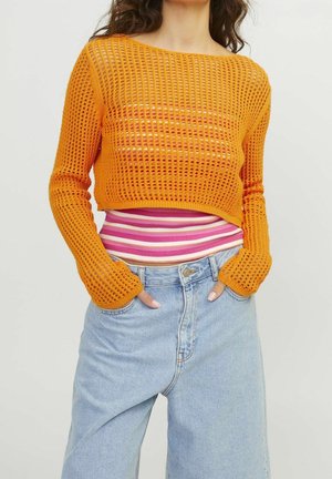 Femme portant un crop top orange en maille à manches longues par-dessus une chemise rayée rose et blanche, avec un jean taille haute bleu clair, mains dans les poches.