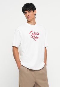 Camiseta de algodón blanca con un logo rojo de "Calvin Klein", de mangas cortas, corte relajado y cuello redondo, combinada con pantalones cortos beiges.