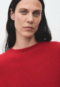 Donna con lunghi capelli scuri e occhi azzurri, che indossa un maglione rosso lavorato a maglia, su uno sfondo chiaro e uniforme.