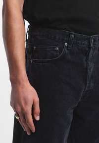 Vaqueros de denim negro con un diseño de pierna recta, cinco bolsillos, remaches en tono plateado y un acabado texturizado. Llevado con una camiseta negra.