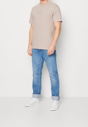 Beiges Baumwoll-T-Shirt mit besticktem Logo, kombiniert mit hellblauen Jeans. Das Model trägt weiße Turnschuhe und hochgekrempelte Hosenbeine.