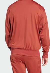 Giacca sportiva rossa con una superficie liscia e opaca, dotata di polsini e orlo elastici. Il design presenta una vestibilità comoda e dettagli di cucitura minimi.