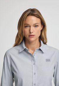 Ljusgrå långärmad skjorta med knappar och krage, med en fyrkantig tonad logotyp på vänster sida och slät textur.