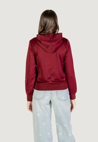 Felpa con cappuccio bordeaux con polsini e orlo a costine. Indossata con jeans blu chiaro caratterizzati da sottili motivi logo sul tessuto.