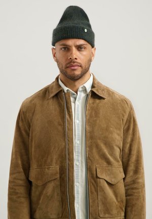 Man met een kort geknipte baard, die een donkergroene gebreide beanie, een beige suède jas over een wit overhemd draagt, kijkt recht vooruit tegen een effen achtergrond.