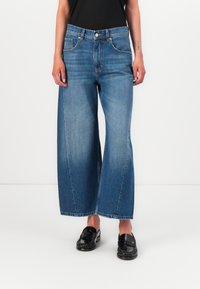 Wide-leg denim jeans in medium blue, featuring a high waist, five-pocket design, and a subtle fade. Paired with black loafers.