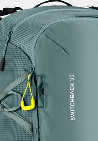 Grüner Stoff-Rucksack mit Gittermuster, ausgestattet mit einem gelben Reißverschlussanhänger, verstellbaren Trägern und weißer Markenbeschriftung "SWITCHBACK 32" an der Seite.