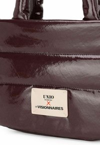 Bolso tote burdeos con acabado brillante, costuras prominentes y una etiqueta con el logo "UNIO HAMBURG X LES VISIONNAIRES" en blanco y negro.
