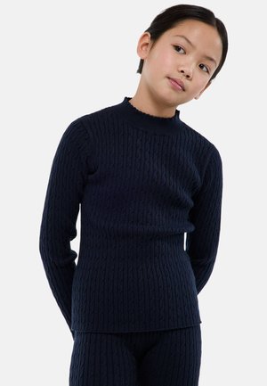 Enfant aux cheveux courts et foncés portant un pull en maille torsadée bleu marine et un pantalon assorti, debout les mains derrière le dos.