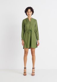 Robe vert olive avec des manches longues, devant boutonné, taille avec cordon ajustable et jupe évasée. Portée avec des talons noirs à lanières.