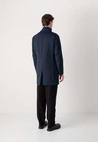 Uomo in piedi, di spalle, che indossa un lungo cappotto blu navy, pantaloni neri e scarpe nere, su uno sfondo chiaro e semplice.