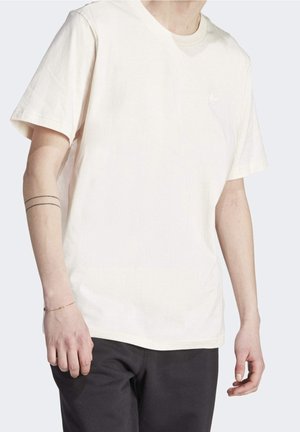 Person trägt ein schlichtes, cremefarbenes kurzärmeliges T-Shirt und schwarze Hosen, mit einem dünnen doppellinienförmigen Tattoo am linken Unterarm.