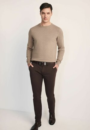 Beżowy sweter robiony na drutach z okrągłym dekoltem, w połączeniu z ciemnobrązowymi spodniami slim-fit i czarnymi skórzanymi butami, tworząc casualowy zestaw.