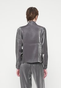 Blusa di un grigio metallico con una leggera lucentezza, spalle strutturate e maniche lunghe. Presenta un colletto e una vestibilità comoda, abbinata a pantaloni coordinati.