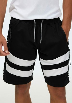 Hombre con pantalones cortos negros con rayas horizontales blancas y cintura con cordón, manos cerca de los bolsillos, combinado con una camisa blanca.