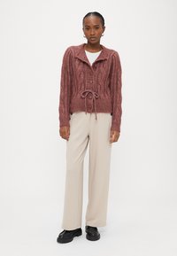 Γυναίκα που φοράει μαρουνένια ζακέτα με κουμπιά και πλεκτό μοτίβο καλωδίωσης, με ζώνη στη μέση, μπεζ παντελόνι με φαρδιά μπατζάκια και μαύρα χοντρά παπούτσια, στέκεται μπροστά σε λευκό φόντο.