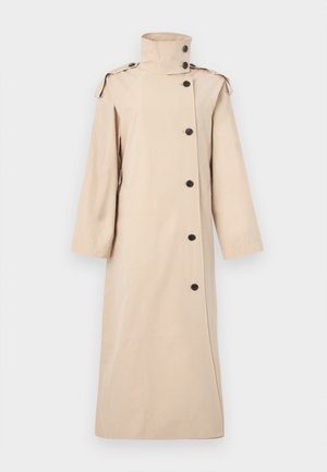 Lange beige trenchcoat met hoge kraag, asymmetrische zwarte knoopsluiting, schouderepauletten en lange mouwen.