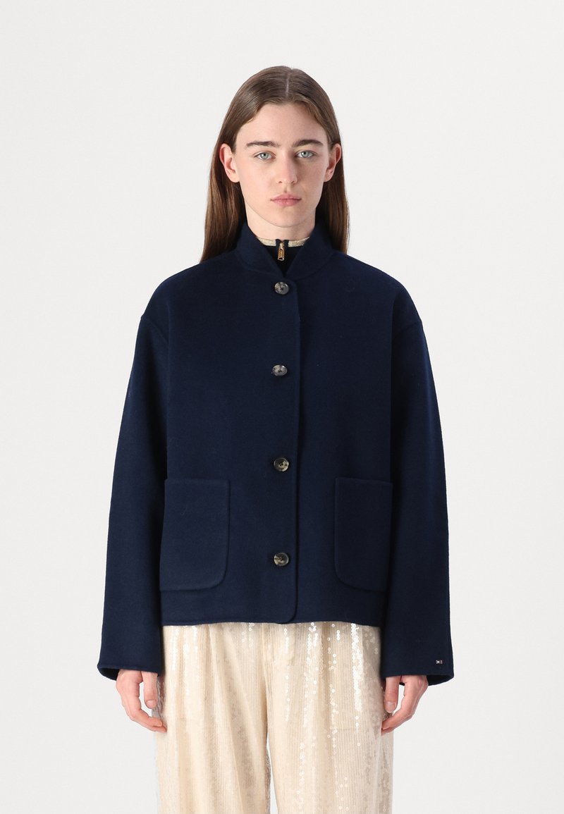 Marineblaues Wolljackett mit hohem Kragen, Knopfschluss und zwei vorderen Taschen. Es hat eine lockere Passform und eine glatte Textur.