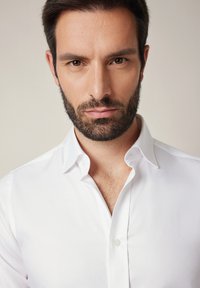 Biała koszula zapinana na guziki z klasycznym kołnierzem, o gładkiej fakturze i dopasowanym kroju. Tkanina wydaje się być świeża i starannie wyprasowana.