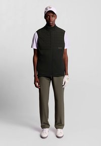 Colete matelassê verde escuro sobre uma camisa branca, combinado com calças verde-oliva e sapatos de golfe brancos. Acessórios incluem um boné e uma luva brancos.