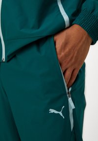 Giacca sportiva teal con accenti azzurri e tasche con zip. Presenta polsini elastici e un logo Puma bianco sulla parte inferiore della gamba.