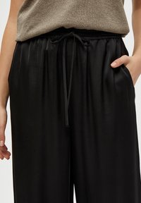 Svarta, vida byxor med elastisk midja, snörning och sidofickor. Sättig, mjuk tyg med lätt glans.